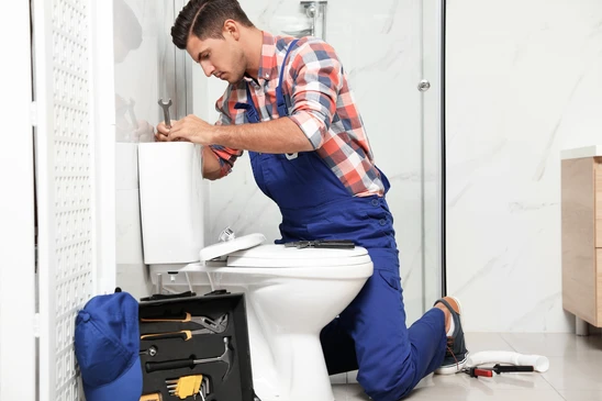 Plombier en salopette bleue et chemise à carreaux intervient dans salle de bains moderne, réparant une cuvette WC suspendue