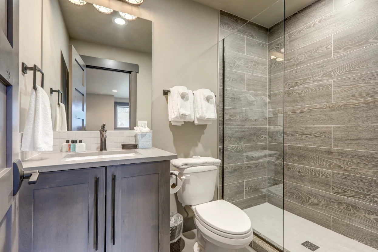Salle de bain moderne avec douche à l'italienne aux carreaux effet bois gris, meuble-vasque en bois foncé avec plan en quartz blanc, miroir encadré noir, toilettes blanches et accessoires coordonnés