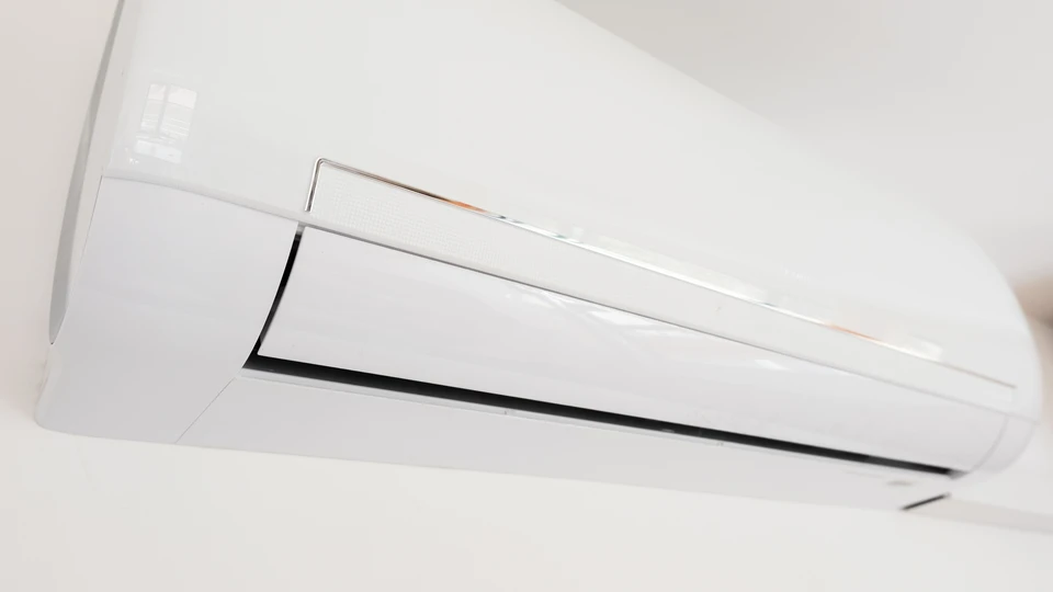 Content que la jeune femme s'installe sur un canapé, tenant une télécommande pour régler son climatiseur fixé au mur, appréciant l'air frais dans un cadre moderne et lumineux du salon